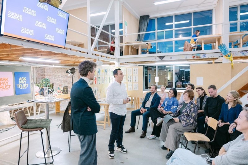 Two men standing, speaking in front of a small audience, indoors